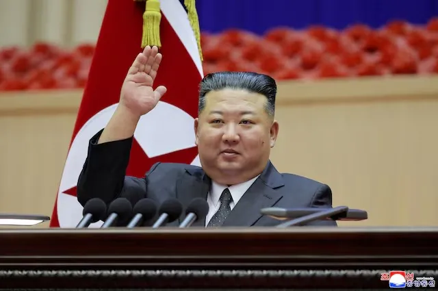 Nhà lãnh đạo Triều Tiên Kim Jong Un mặc vest đen đang giơ tay phát biểu trước bục, phía sau là quốc kỳ Triều Tiên - Ảnh 1.